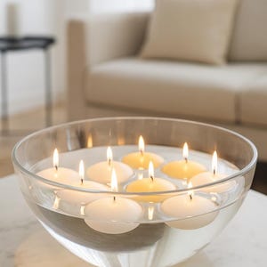 Op de afbeelding: Een glazen kom gevuld met water en meerdere drijvende, brandende kaarsen. De ronde kaarsen geven een warm licht. De kom staat op een wit marmeren oppervlak, met een bank in neutrale tinten op de achtergrond.