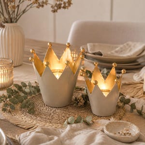 Op de afbeelding: Twee kroonvormige kaarsenhouders in verschillende maten, met een matte gebroken witte buitenkant en een metallic gouden binnenkant. Elk punt van de kroon is afgewerkt met een klein gouden bolletje en ze bevatten brandende theelichtjes.