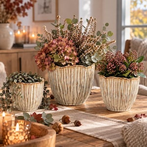Può includere: Tre vasi in ceramica con una texture a coste verticali e una smaltatura color crema con accenti marroni lungo le creste. Ogni vaso presenta un bordo smerlato e contiene un diverso arrangiamento di fogliame verde e marrone rossastro.