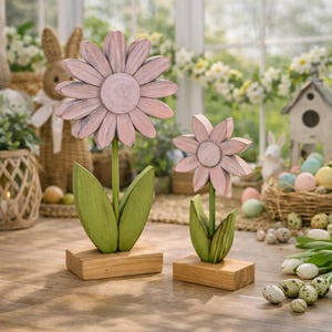 Könnte beinhalten: Zwei Holzblumen-Dekorationen. Die Blumen sind hellrosa mit grünen Stielen und Blättern, auf Holzblöcken. Der Hintergrund zeigt Osterdekorationen wie Eier und einen Hasen.