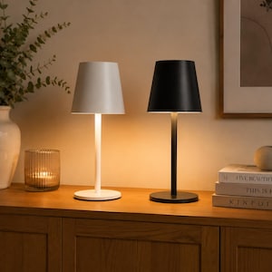Könnte beinhalten: Zwei kabellose Tischleuchten in schlichtem, modernem Design. Eine Leuchte hat eine mattweiße Oberfläche, die andere eine mattschwarze Oberfläche. Beide Leuchten bestehen aus einem schlanken zylindrischen Stiel, einem runden Fuß und einem konischen Schirm.