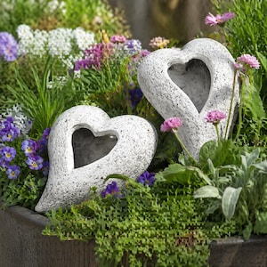 Könnte beinhalten: Zwei strukturierte, weiße herzförmige Gartenornamente mit herzförmigen Ausschnitten, eingebettet in lebendiges grünes Laub und bunte Blumen. Die Ornamente verleihen dem Garten eine romantische Note.