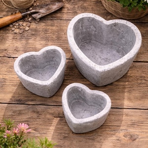 Pode incluir: Três vasos cinzentos em forma de coração de tamanhos variados sobre uma superfície de madeira. Os vasos têm uma aparência texturizada e estão vazios. Luvas de jardinagem, uma espátula e barbante são visíveis no fundo.