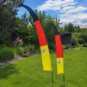 Könnte beinhalten: Zwei Fußball-Außenfahnen mit schwarz-rot-gelbem Design und einem Fußball-Motiv. Die Fahnen sind auf einer grünen Wiese unter blauem Himmel mit Wolken aufgestellt. Die Fahnen sind aus Stoff.