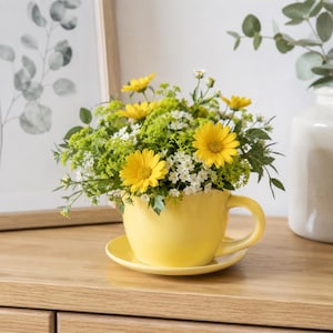 Puede incluir: Una taza y platillo amarillos llenos de un vibrante arreglo de margaritas amarillas, flores blancas y follaje verde. El conjunto se coloca sobre una superficie de madera, creando una presentación alegre.