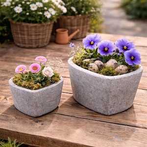 May include: Two heart-shaped gray stone planters filled with colorful flowers and moss sit on a wooden table. One planter has pink and white flowers, the other has purple flowers and small speckled eggs.