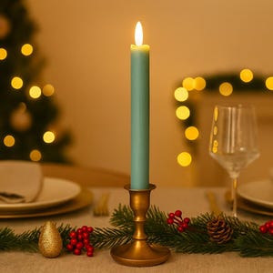 May include: A lit teal candle in a gold candlestick sits on a table set for a holiday meal. The table is decorated with a garland of greenery, red berries, and a gold ornament. A Christmas tree with bokeh lights is in the background.