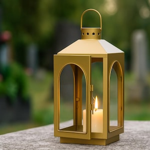 May include: A gold metal lantern with a lit candle inside. The lantern has a handle on top and a door that is slightly ajar. The background is blurred, showing a cemetery with headstones and greenery.