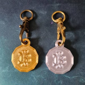 May include: Two gold-colored keychains with a gold-colored metal clasp. Each keychain has a round, octagonal-shaped charm with the letter "B" embossed on it. One charm is gold-colored and the other is silver-colored.