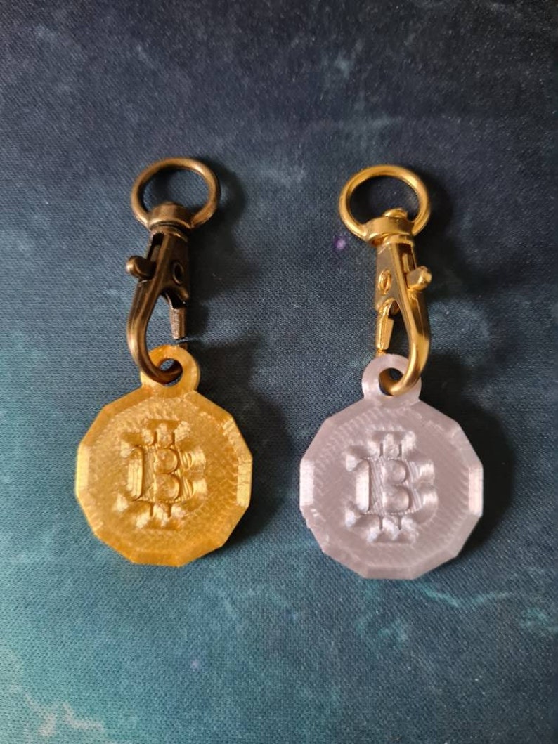 May include: Two gold-colored keychains with a gold-colored metal clasp. Each keychain has a round, octagonal-shaped charm with the letter "B" embossed on it. One charm is gold-colored and the other is silver-colored.