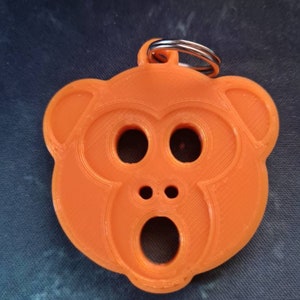 以下が含まれることがあります： オレンジ色のプラスチック製のキーホルダーで、大きな口を開けた猿の顔の形をしています。目は小さく2つあります。キーホルダーには銀色の金属製のリングが付いています。