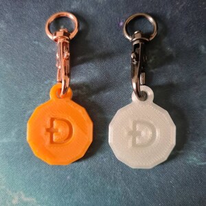 May include: Two 3D printed keychain charms featuring a stylized Bitcoin symbol. One charm is orange and the other is white. Both charms have a silver metal clasp.