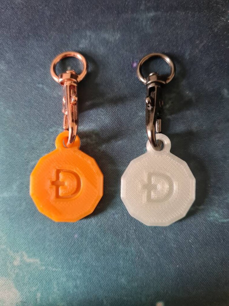 May include: Two 3D printed keychain charms featuring a stylized Bitcoin symbol. One charm is orange and the other is white. Both charms have a silver metal clasp.