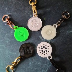 May include: Five different cryptocurrency keychains with various logos. The keychains are made of different materials, including metal, plastic, and 3D printed plastic. The logos include Bitcoin, Ethereum, Cardano, and a logo that looks like a circle with a dot in the center.