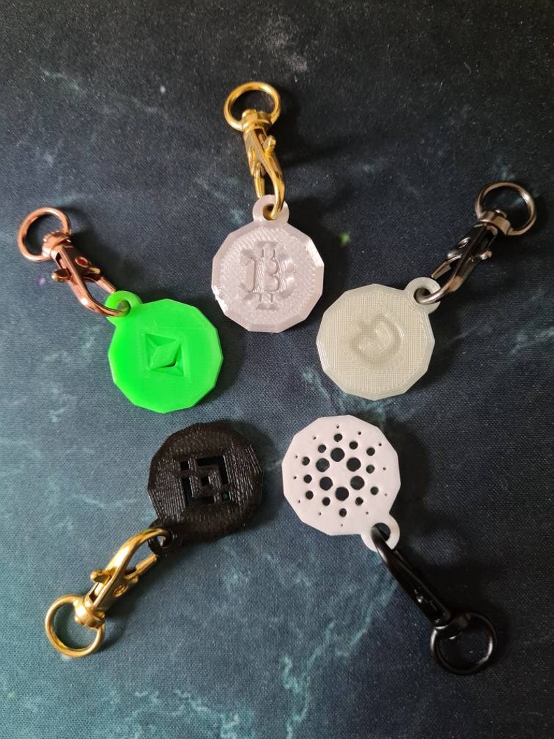May include: Five different cryptocurrency keychains with various logos. The keychains are made of different materials, including metal, plastic, and 3D printed plastic. The logos include Bitcoin, Ethereum, Cardano, and a logo that looks like a circle with a dot in the center.