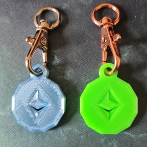 May include: Two keychains with a silver and a rose gold clasp. Each keychain has a round, flat charm with a raised Ethereum logo. One charm is blue and the other is green.