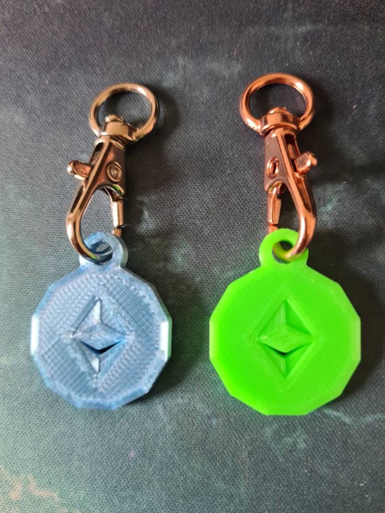 May include: Two keychains with a silver and a rose gold clasp. Each keychain has a round, flat charm with a raised Ethereum logo. One charm is blue and the other is green.