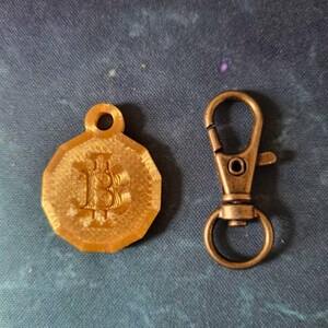 May include: A gold-colored keychain with a Bitcoin symbol on a round charm and a bronze-colored metal clasp.