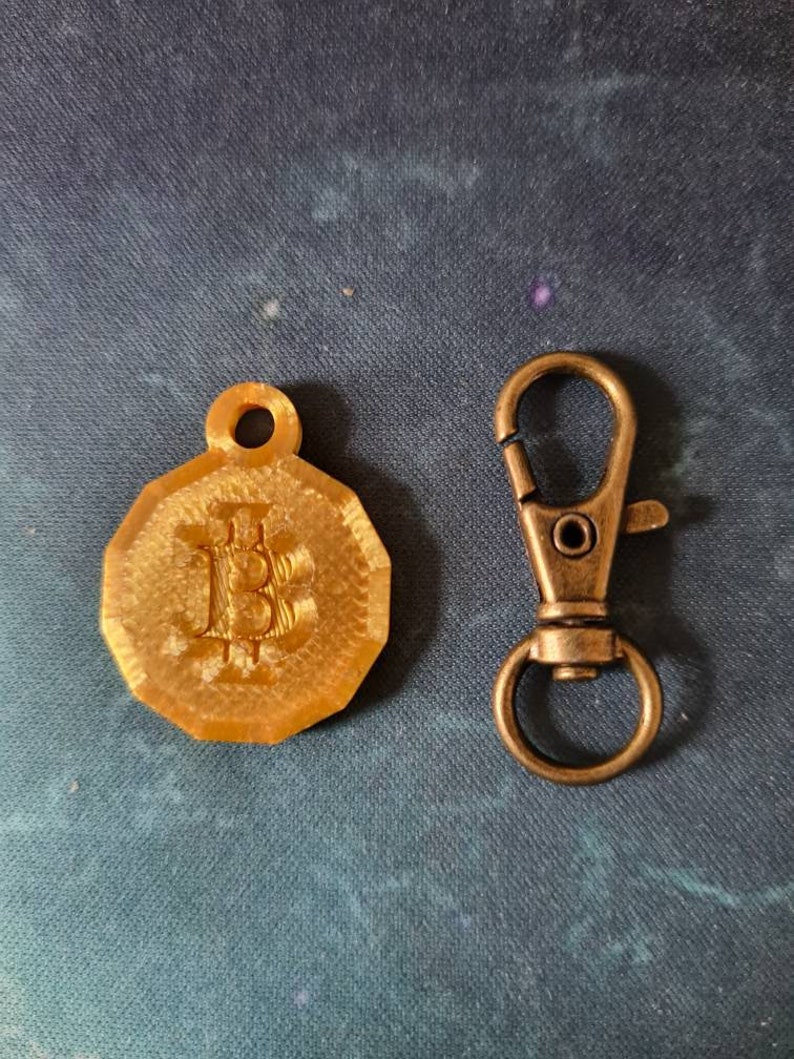 May include: A gold-colored keychain with a Bitcoin symbol on a round charm and a bronze-colored metal clasp.
