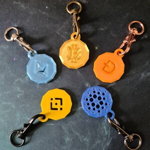 May include: Five different cryptocurrency keychains with metal clasps. The keychains are round and feature different cryptocurrency logos. The logos are printed in blue, orange, and yellow.