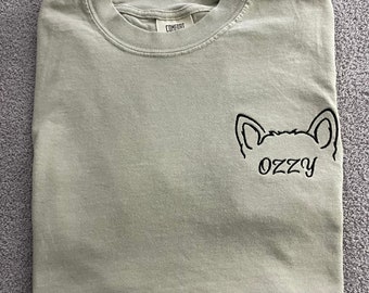 May include: A sage green t-shirt with a black embroidered cartoon dog and the word "OzZy" on the left chest.