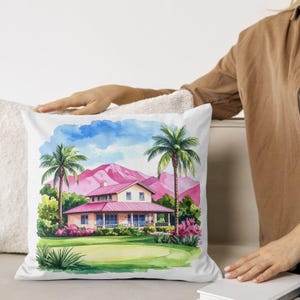 May include: White throw pillow with a watercolor painting of a house with a pink roof, palm trees, and pink mountains under a blue sky. The pillow has a green lawn in the foreground.