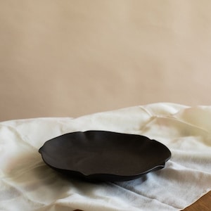 May include: A black ceramic serving dish with a wavy edge. The dish is sitting on a white tablecloth.
