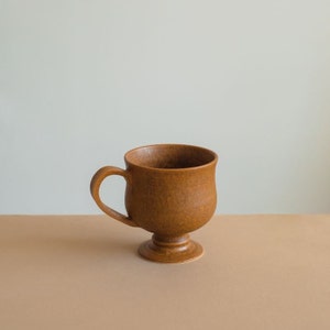 May include: A brown ceramic mug with a handle and a pedestal base. The mug has a textured surface.