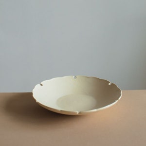 May include: A white ceramic serving bowl with a scalloped edge and small cutouts. The bowl is sitting on a light brown surface.