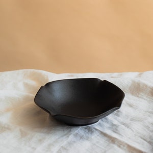 Op de afbeelding: Een zwarte keramische kom met een golvende, bloemblaadjes-achtige rand. De kom staat op een wit linnen tafelkleed.