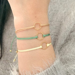 May include: Three delicate bracelets with gold circle charms. The bracelets are made of thin, braided cords in beige, green, and white.