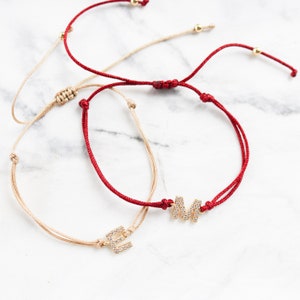 Peut inclure: Deux bracelets ajustables en cordon avec des breloques en lettres dorées. Le bracelet de gauche a un charme "E" en or et le bracelet de droite a un charme "M" en or. Les deux breloques sont serties de pierres de zircone cubique transparentes.