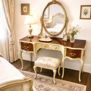 May include: A vintage vanity table with a gold-framed oval mirror, a small floral stool, and a decorative lamp. The table has a dark brown top with cream-colored accents and ornate drawer pulls. A vase of flowers and framed art complete the elegant bedroom scene.