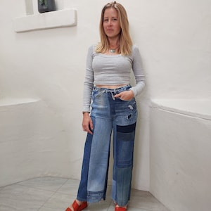 Op de afbeelding: Een vrouw draagt een grijs, lang mouwig, getailleerd topje met een blauwe patchwork broek met wijde pijpen. Ze draagt een zilveren ketting en heeft lang, bruin haar.