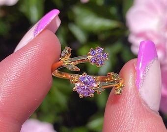 May include: A gold ring with a delicate floral design featuring purple and clear gemstones. The ring is adjustable and has a vine-like stem.