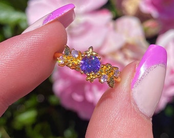 May include: A gold ring with a purple gemstone surrounded by smaller clear and purple stones. The ring has a delicate floral design.