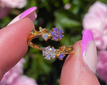 May include: Gold ring with a delicate design featuring a butterfly and two clusters of purple and clear cubic zirconia stones. The ring is styled as a floral motif.