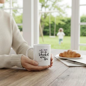 Könnte beinhalten: Weiße Keramik-Tasse mit schwarzem Schriftzug: "ich muss los, die Schaukel wird frei." Ein Croissant liegt auf einem Teller neben der Tasse. Eine Person hält die Tasse vor einem Fenster mit einer Schaukel und einem Kind im Hintergrund.
