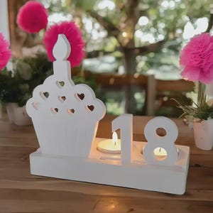 Könnte beinhalten: Weißer Kerzenhalter aus Holz in Cupcake-Form mit Herz-Ausschnitten und einer kerzenförmigen Flamme. Die Zahl "18" wird neben dem Cupcake mit zwei brennenden Kerzen angezeigt. Der Hintergrund umfasst rosa Pompons und Grün.