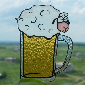 May include: A stained glass beer mug with a sheep's head on top, hanging from a suction cup. The mug is filled with yellow liquid and topped with white foam. The background shows a green landscape and a blue sky.