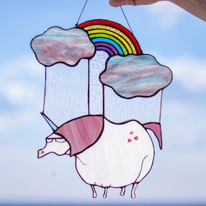 Può includere: Un acchiappasole in vetro colorato raffigurante un unicorno bianco con criniera rosa e tre piccoli cuori rossi sul fianco. Sopra l'unicorno ci sono due nuvole iridescenti collegate da un arcobaleno colorato, con strisce di pioggia in vetro.
