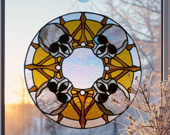 May include: A stained glass window hanging in a window with a white frame. The window is round and features a design of four skulls in a circle with a yellow background and brown accents.