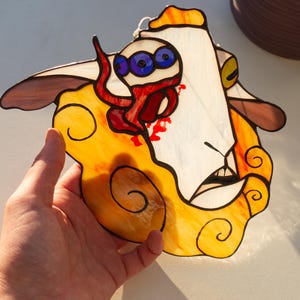 May include: Stained glass artwork featuring a surreal face with three blue eyes, red tentacles, and a yellow and orange cloud-like background. The artwork has brown and white accents and is held by a hand.