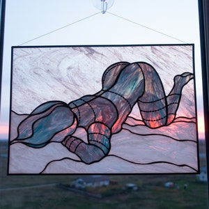 May include: A stained glass window hanging in a window frame. The stained glass depicts a person in a kneeling position with their head resting on their hand. The stained glass is made of various shades of blue, green, red, and orange glass.