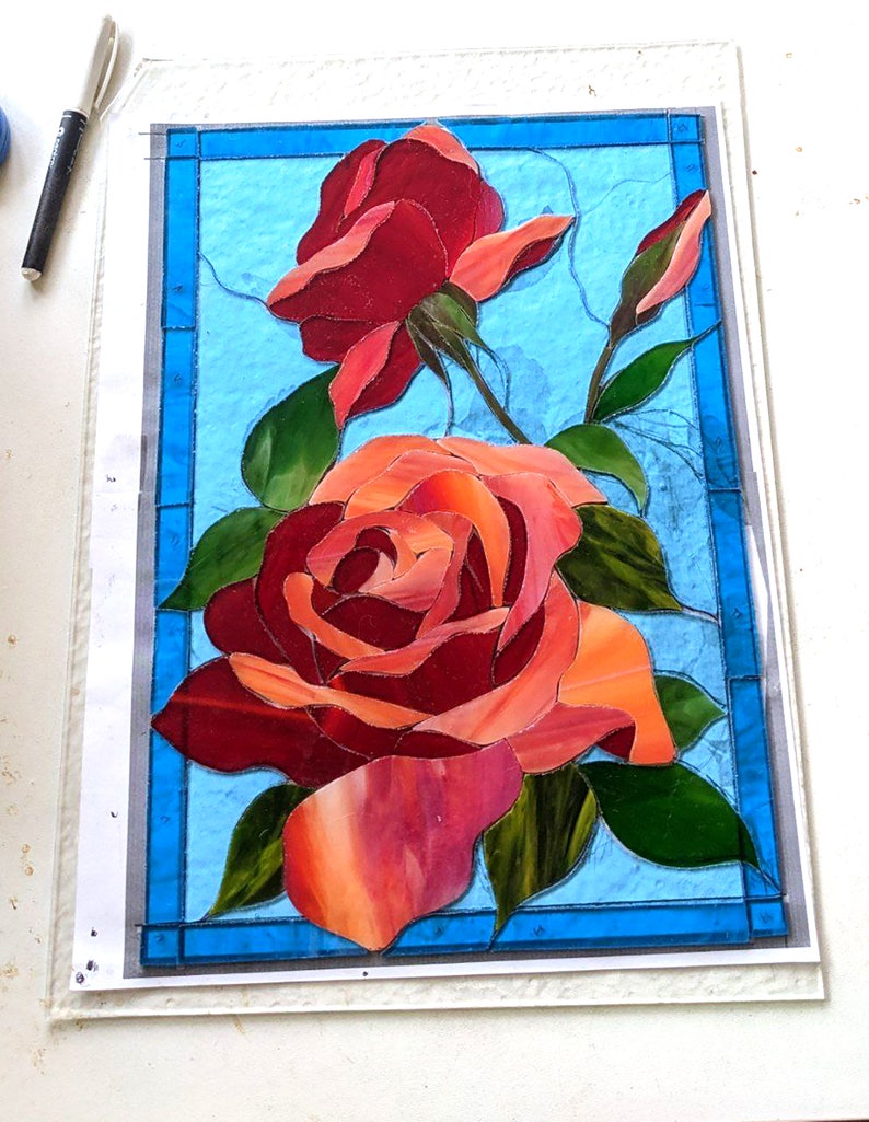 May include: A stained glass artwork featuring two red roses with green leaves on a blue background.