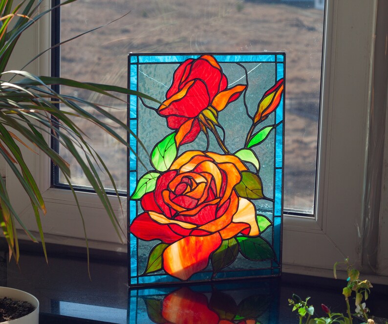 May include: A stained glass window panel featuring two red and orange roses with green leaves and stems. The panel is framed in a blue border.