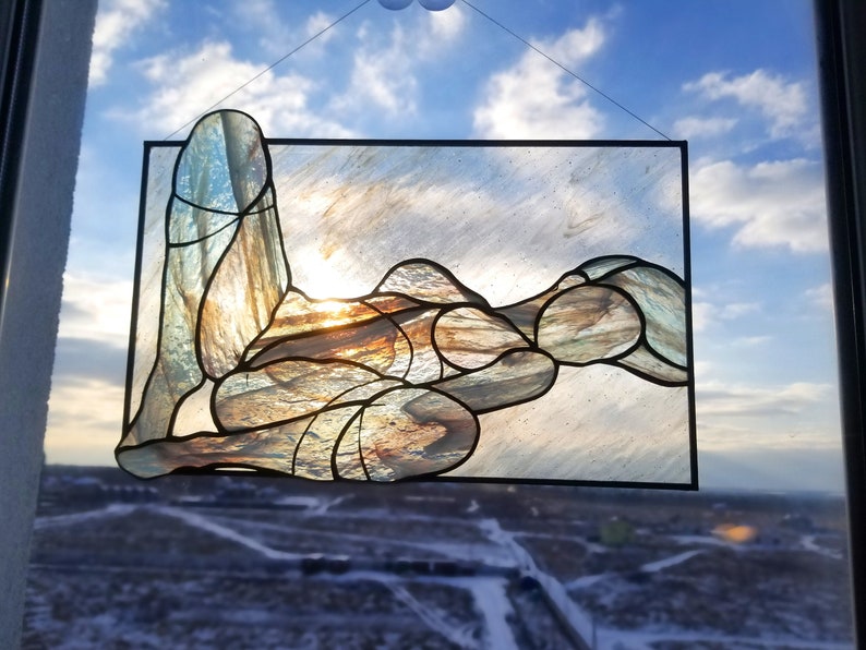 May include: A stained glass window depicting a stylized figure in a reclining pose. The figure is made of various shades of blue, brown, and gold glass. The background is a cloudy sky.
