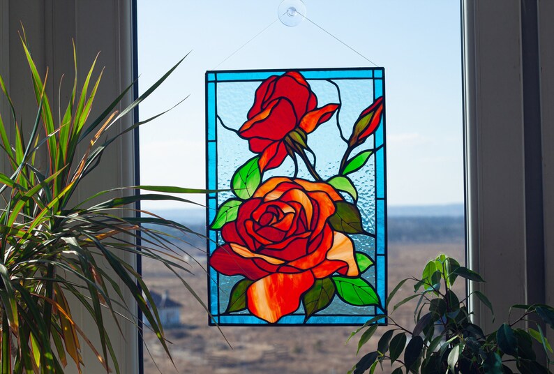 May include: A stained glass window panel featuring three red roses with green leaves and stems. The panel is framed in a blue border and hangs in a window with a view of a field.
