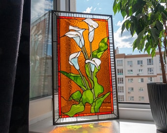 May include: Stained glass panel featuring white calla lilies with green leaves against an orange background. The artwork is framed in black and has a red border. The panel is displayed on a windowsill.