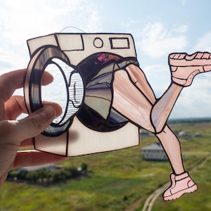 May include: A stained glass art piece depicting a washing machine with legs and feet. The legs and feet are pink, and the washing machine is white with a black and clear glass door. The artwork is set against a backdrop of a green field and blue sky.